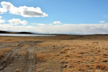 A field gravel road descends from the yellowed hills to the shore of a large lake in the mountains under a cloudy autumn sky.