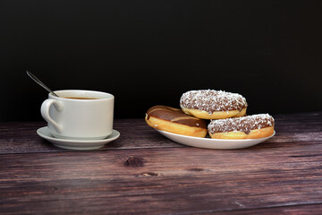 A plate with several donuts in chocolate icing and a cup of hot coffee on a saucer with a spoon on a wooden table.