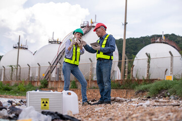 Environmental scientists or workers, wearing safety helmets and gloves, examining waste materials along a shoreline are large industrial tanks environmental waste water or pollution effects concept.