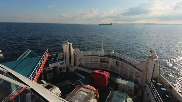 A large ferry cruises through open waters under a clear sky, offering expansive ocean views. Vehicles are parked on deck, and another ship is visible in the distance