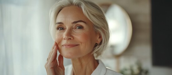Close-up portrait of a beautiful mature woman with gray hair, looking at the camera with a gentle smile, touching her face with her hand. - obrazy, fototapety, plakaty