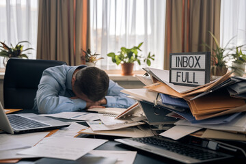 An employee slumped over a desk, overwhelmed by work, with an overflowing inbox