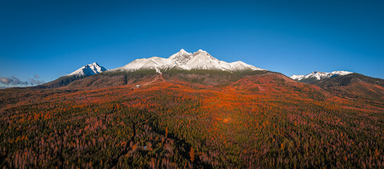 Fototapeta premium Tatranske Matliare, Slovakia - Aerial view of the snowy mountains of Lomnicky Peak in the High Tatras with beautiful red and orange coloured autumn trees and foliage and clear blue sky at Vysoke Tatry