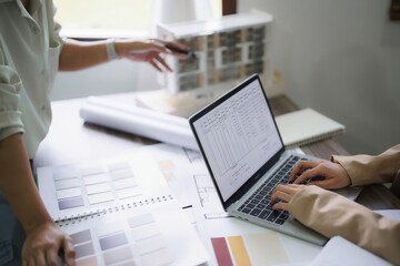 Female architects explain building model and interior design to partner while typing data on laptop