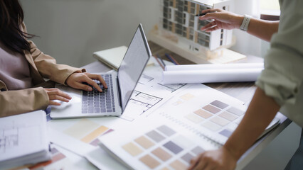 Female architects explain building model and interior design to partner while typing data on laptop
