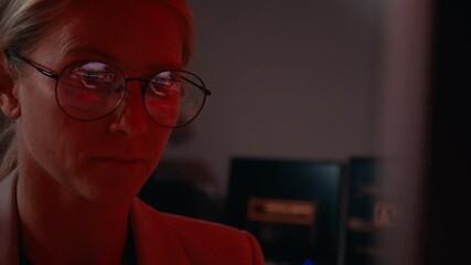Closeup of face of stressed Caucasian female IT security specialist, red flashing warning light reflecting on glasses, frantically working to defend system from firewall breach or hacking attack