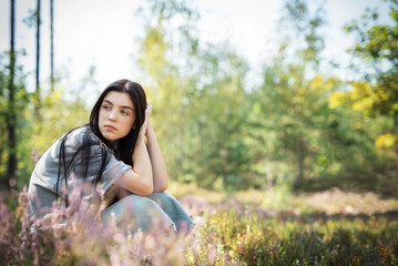 A young woman sitting thoughtfully in a serene forest clearing surrounded by colorful wildflowers on a sunny day