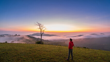 A man stands on a grassy hill overlooking a beautiful landscape. The sky is a mix of orange and blue, and the clouds are white and fluffy. The man is wearing a red jacket and he is enjoying the view