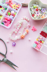 Crafting colorful beaded jewelry on a pastel pink workspace with scissors and various charms displayed in containers