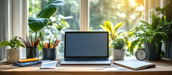 Fototapeta premium A laptop with a blank screen sits on a wooden desk with a window in the background, along with houseplants, notebooks, pencils, and a clock.