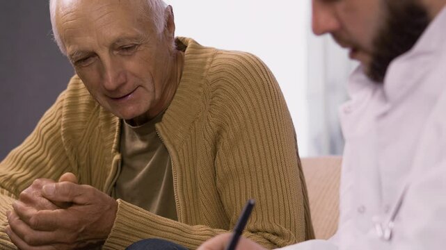 Senior man talking to a doctor while being in a home visit, speak about his health and problem sitting on sofa.