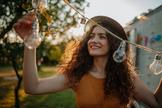 Young beautiful woman is on a camping trip in nature, putting lights on tree, preparing for evening.