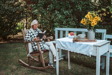 Beautiful older women is sitting in rocking chair in the garden and reading book. Female senior is...