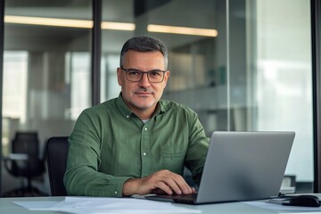 Fototapeta premium Middle-aged man with glasses working on a laptop at an office desk, blurred glass wall background, wide-angle professional photograph.
