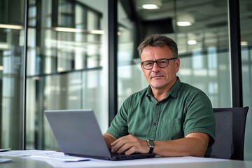Fototapeta premium Middle-aged man with glasses working on a laptop at an office desk, blurred glass wall background, wide-angle professional photograph.