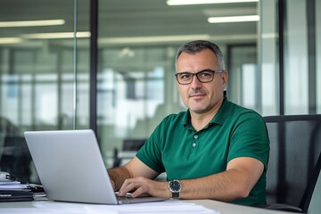 Fototapeta premium Middle-aged man with glasses working on a laptop at an office desk, blurred glass wall background, wide-angle professional photograph.