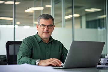 Fototapeta premium Middle-aged man with glasses working on a laptop at an office desk, blurred glass wall background, wide-angle professional photograph.