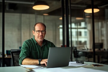 Fototapeta premium Middle-aged man with glasses working on a laptop at an office desk, blurred glass wall background, wide-angle professional photograph.