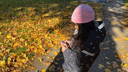 Beautiful Indonesian little girl holding mini kid camera in vibrant autumn season wearing padded jacket and pink beanie. Figure viewed from behind