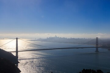 Obraz premium Bright sun reflects on bay behind tower of Golden Gate Bridge with city in distance