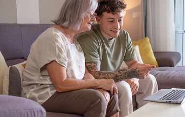 Happy young nephew teaching technology to his old grandmother using laptop. Senior woman learns to...