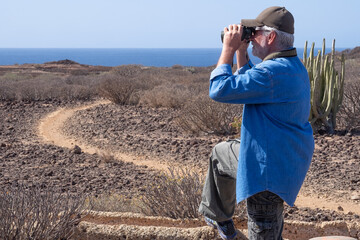 Active happy senior bearded man walking outdoor in a sunny day along a footpath using a binoculars enjoying healthy lifestyle in vacation. Arid landscape, horizon over water