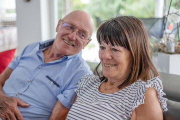 Close up portrait of senior couple sitting together on sofa in the living room with a large window at the back, two serene elderly retiree enjoying a peaceful moment of peace in their home