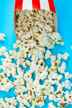 Vertical image of fallen red and white cardboard bucket scattering popcorn on blue background, cinema food concept