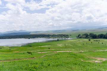 Obraz premium Green fields, reservoir. Ripples on the water. Coniferous and deciduous forest, mountains, sky with clouds in the background