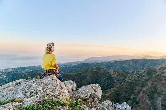 Mature woman wearing wireless headphones and sitting in front of mount Etna, Italy