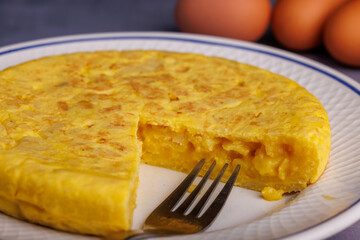 Spanish potato omelette with a missing slice, surrounded by bread, red checkered napkin, and eggs, on a blueish concrete table