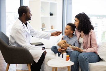 African male doctor measures temperature of young boy while seated on hospital bed. Boy holds teddy bear, accompanied by concerned African mother holding smartphone.