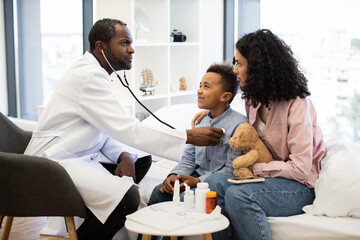 Obraz premium African doctor listens to heartbeat of young African boy with stethoscope. Mother sits beside child offering comfort and holding teddy bear. Scene conveys healthcare, nurturing, and care.