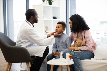 African male doctor examines sick African boy's lymph nodes while mother sits nearby with teddy bear. Medical consultation takes place in bright healthcare setting, promoting care and diagnosis.