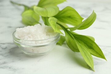 Sea salt in glass bowl and green leaves on white marble table, closeup