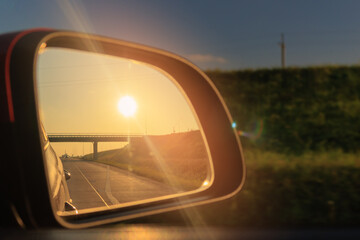 Obraz premium The sun is reflected in the mirror, evening road. View in the side rear view mirror of a red car driving along the highway
