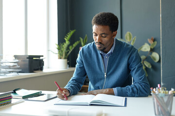 Portrait of African American male teacher sitting at desk in school classroom and taking notes preparing for lesson copy space