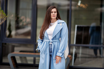 Fototapeta premium Portrait stylish plus size woman with long hair outdoors, wears fashionable light blue trench coat, showcasing trendy and elegant look in urban setting