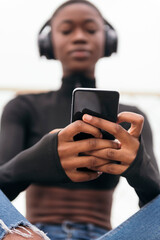 low angle of unrecognizable young glad ethnic female in wireless headphones surfing internet on cellphone while listening to song in city, selective focus on the phone