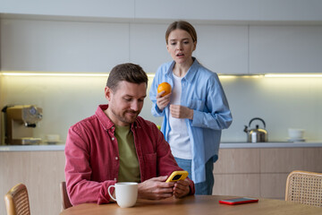 Woman secretly reads husband messages over shoulder while he chats on kitchen. Suspicion, trust...