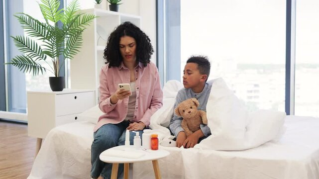 Concerned African mother checks temperature of her sick son while holding phone for online medical consultation. Young boy, wrapped in blanket, clutches teddy bear.