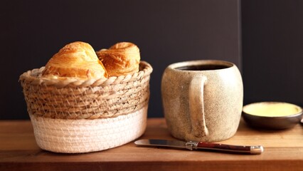 Rustic basket filled with croissants beside ceramic mug and butter on wooden board promotes simple, artisanal breakfast. Focus on flaky pastries paired with rich coffee for cozy meal.