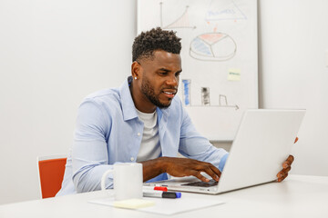 Young man working on a laptop at a desk with a whiteboard in an office during the day