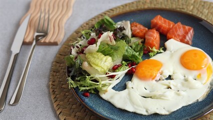 Sunny eggs with bright orange yolks sit next to green salad, pomegranate seeds, and salmon on blue plate, emphasizing vibrant eco-conscious healthy lifestyle meal.