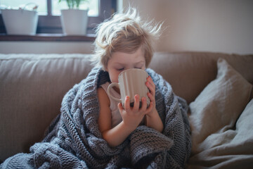 Little boy drinking tea from cup. Sick boy with cold is sitting on sofa under warm blanket and...