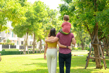 Fototapeta premium Back view of father, mother with little child in the park on a sunny summer day together. Son sitting on his father shoulders happy in vacation time. Happy Family.