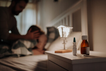 Close up on medication on bedside table. Sick girl with cold lying in the bed, father taking care of her.