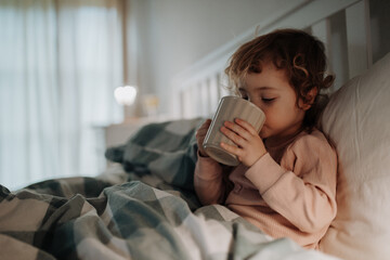 Little girl lying in the bed, coughing and holding tea cup. Sick girl with cold is drinking herbal tea.