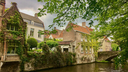 Obraz premium Charming medieval houses line a tranquil canal surrounded by lush greenery in Bruges, Belgium, evoking a sense of peaceful nostalgia and European holiday allure