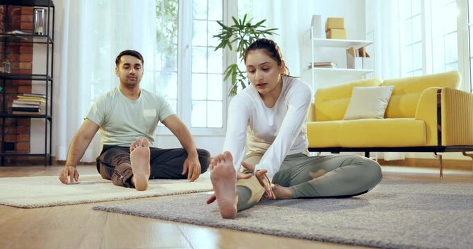 Indian Asian young couple learning yoga in the comfort of their lavish home, performing janu shirasana or head to knee pose together, enhancing flexibility and mindfulness in a serene environment